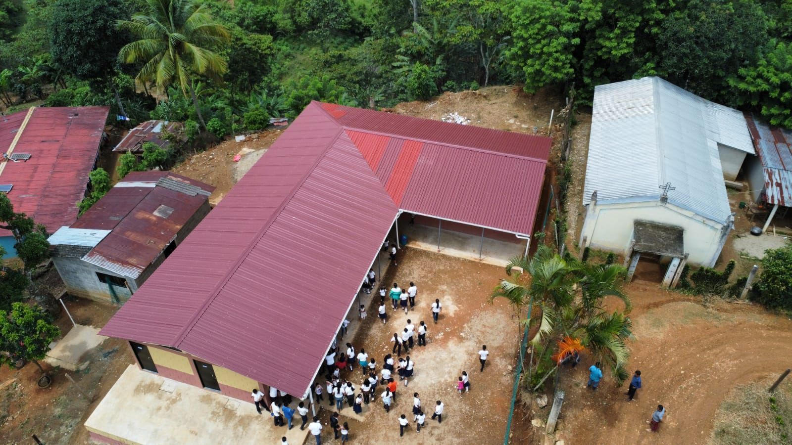 Aerial view of a red-roofed school building with people gathered in the courtyard, surrounded by trees.
