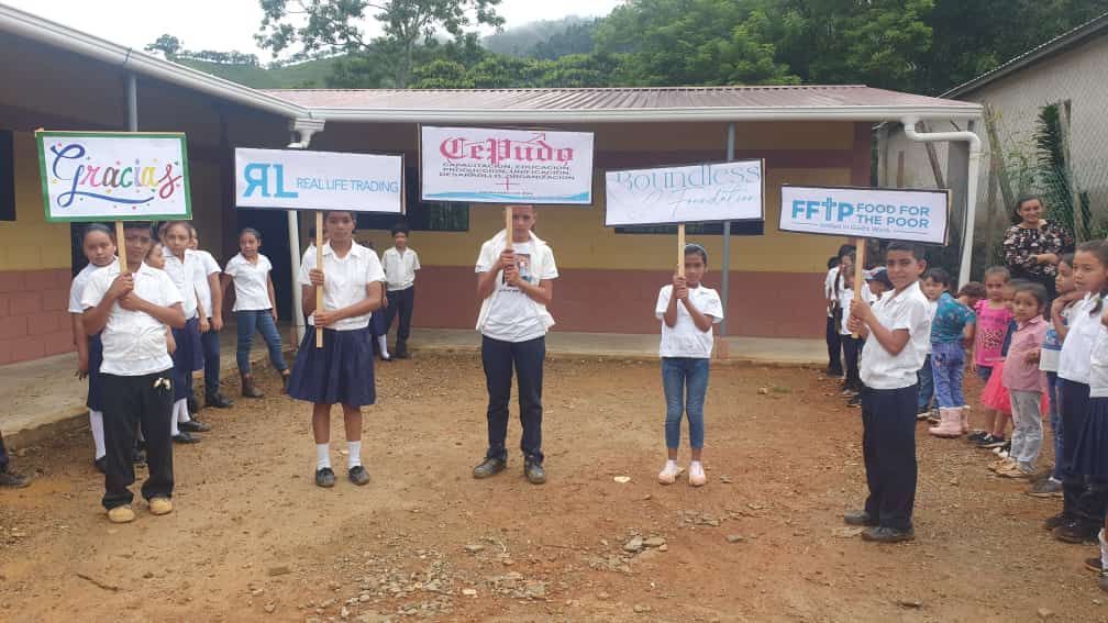 Students stand in a row outdoors holding signs in front of a school building.