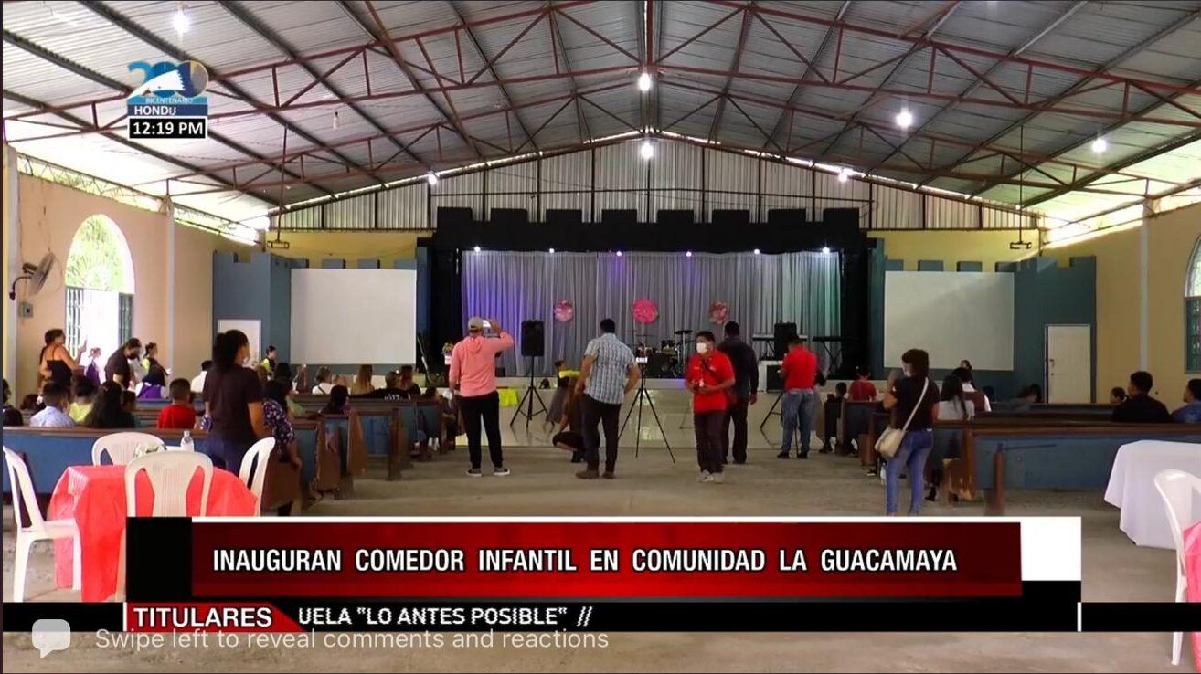 Indoor gymnasium with people gathered near a stage for a community event, banners and screens visible