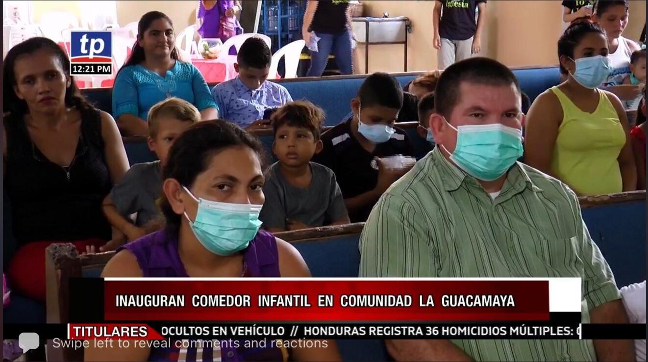 Masked audience seated in a crowded indoor event, with a news-style banner overlay at the bottom.