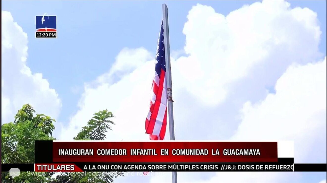 Large U.S. flag on a flagpole against a bright cloudy sky, with a Spanish news banner below
