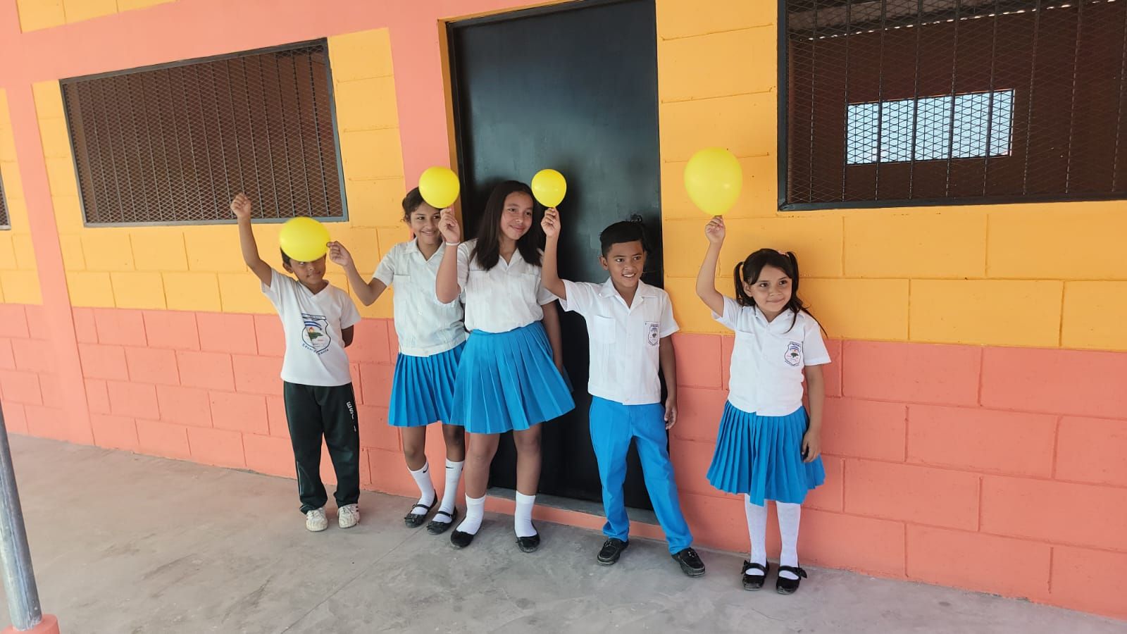 Five students in uniforms stand smiling outside a peach-colored school building, holding yellow balloons.