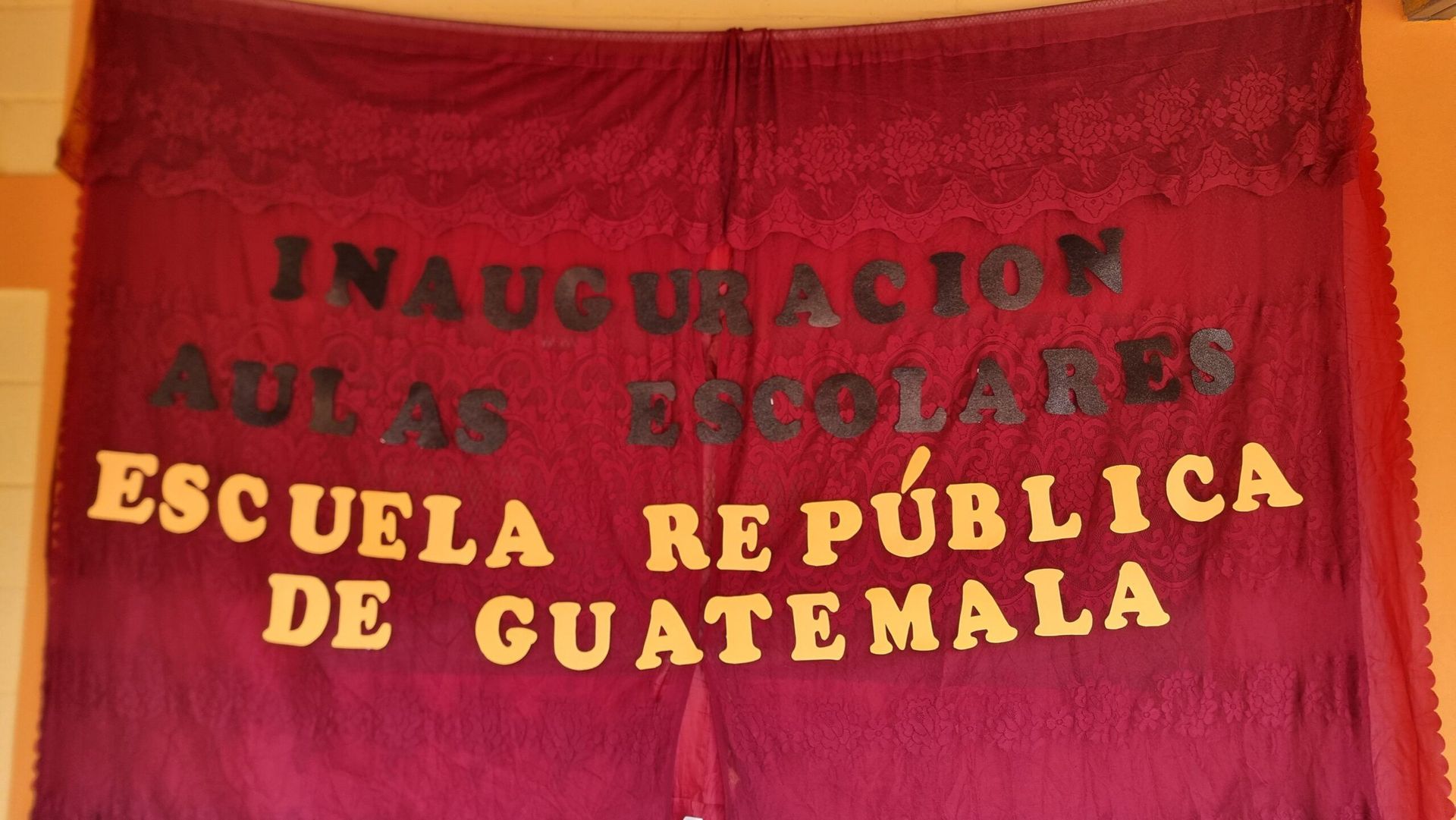 Red curtain with gold text reading “Inauguración Aulas Escolares, Escuela República de Guatemala”