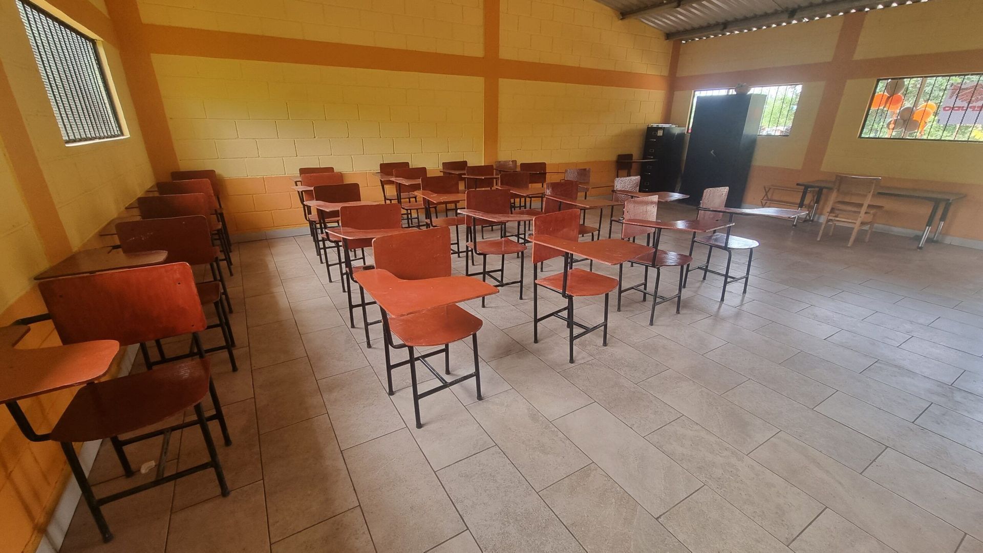 Empty classroom with rows of orange chairs and desks on a tiled floor, lit by windows.