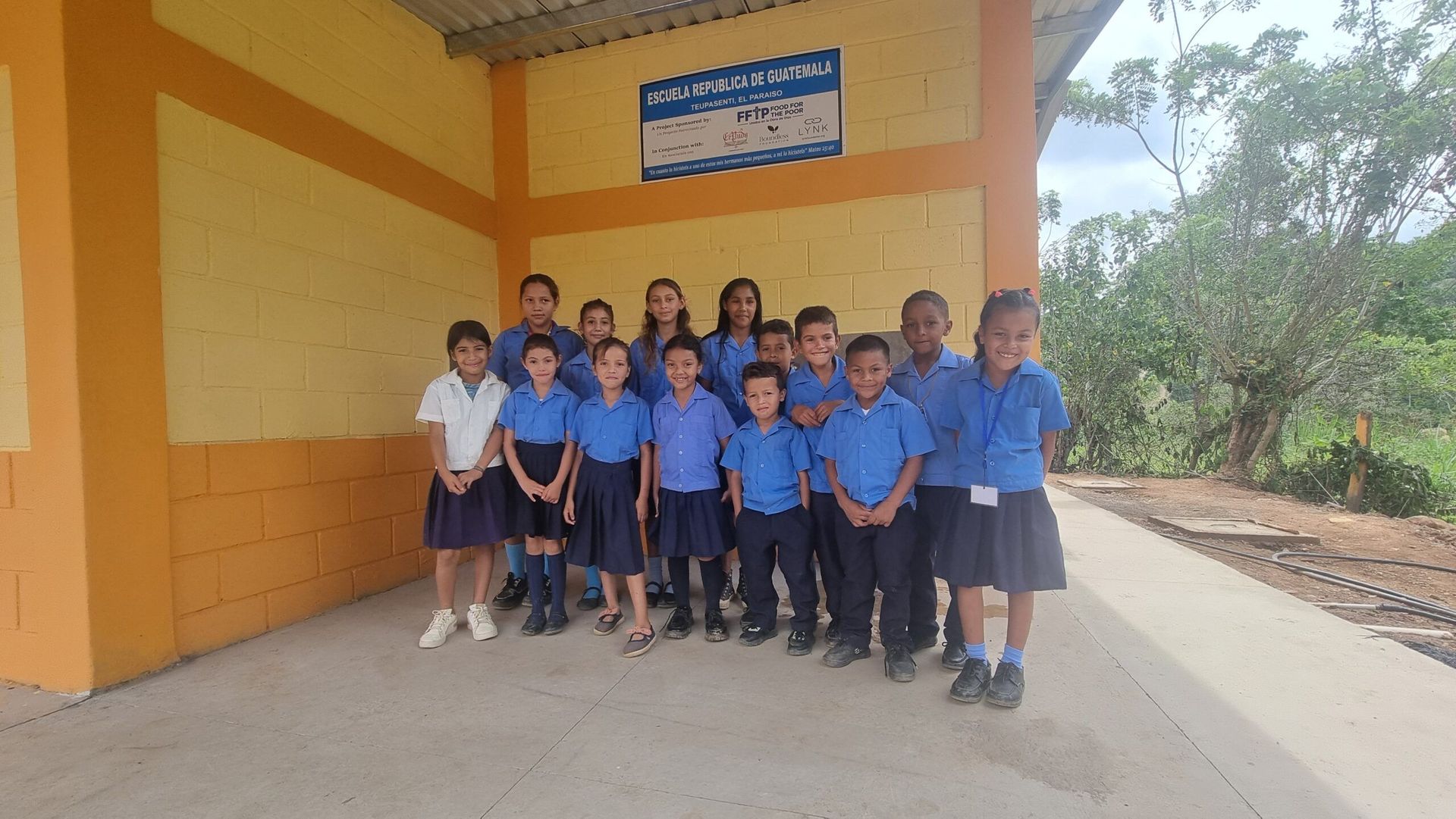 Group of students in blue uniforms posing outside a yellow school building.