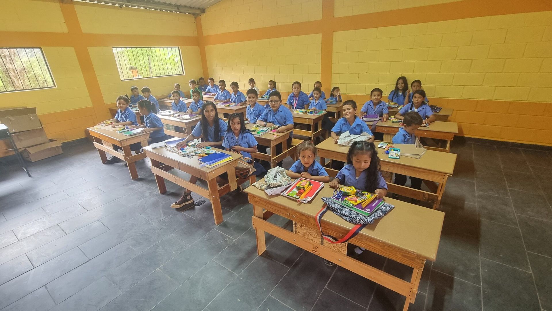 Students seated at desks in a classroom with yellow walls and windows, looking toward the front.