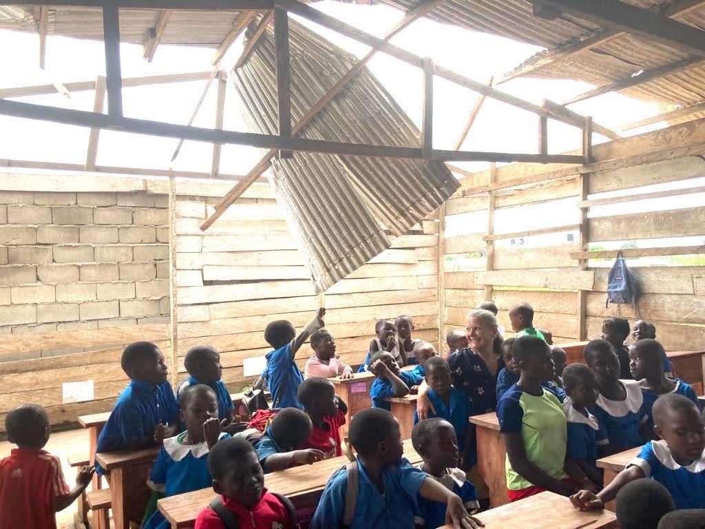 Children seated in a rustic classroom, raising hands during a lesson under a damaged roof.