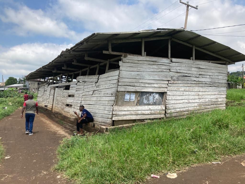 Small white weathered building beside a dirt path, with two people nearby and grassy surroundings.
