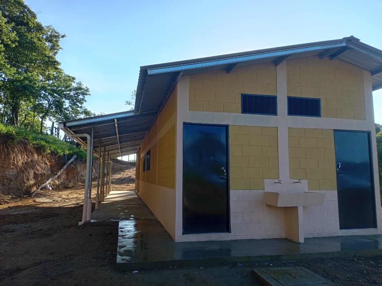 Small yellow building with blue roof and doors on a dirt hillside, viewed from the front.