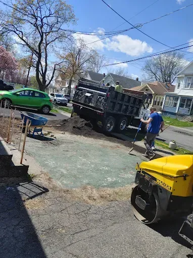 Workers pour a concrete driveway beside a black dump truck, green car, and yellow generator in a neighborhood.