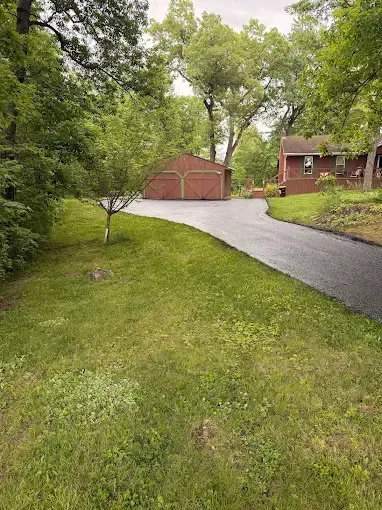 Wet driveway leading to a red garage beside a brick house, surrounded by green trees and lawn