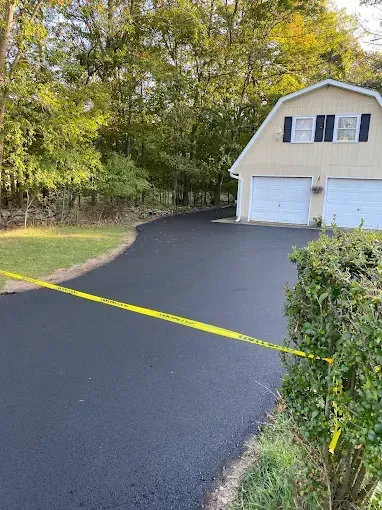 Freshly paved black driveway with yellow caution tape leading to a white garage beside trees.