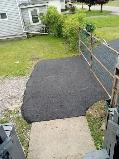 Backyard walkway with a fresh black asphalt patch leading from a concrete step to a chain-link fence gate