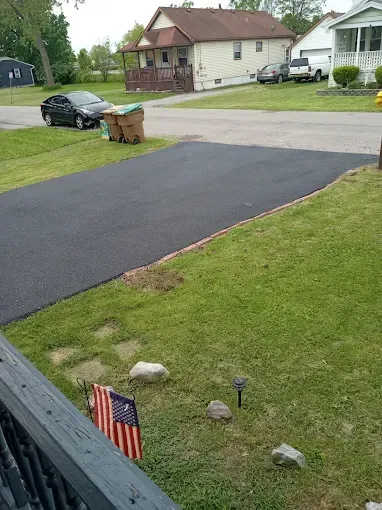 Freshly paved black driveway beside a lawn, with a small American flag in the foreground and houses across the street