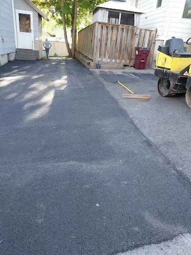 Freshly paved driveway beside a house, with a wooden fence, red bin, and yellow machinery on the right.