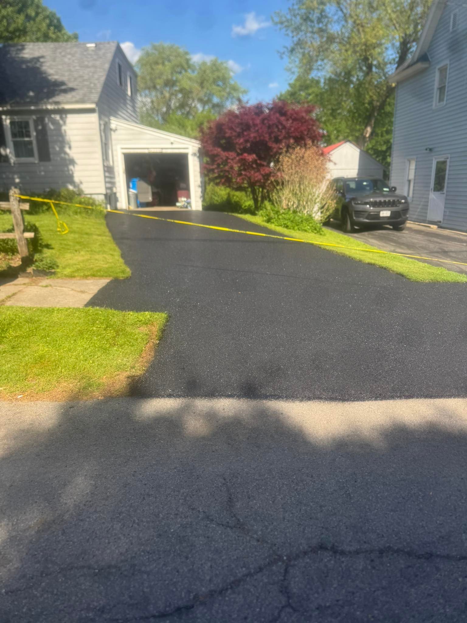 Freshly paved driveway leading to a garage beside houses, with green lawn and a flowering tree.