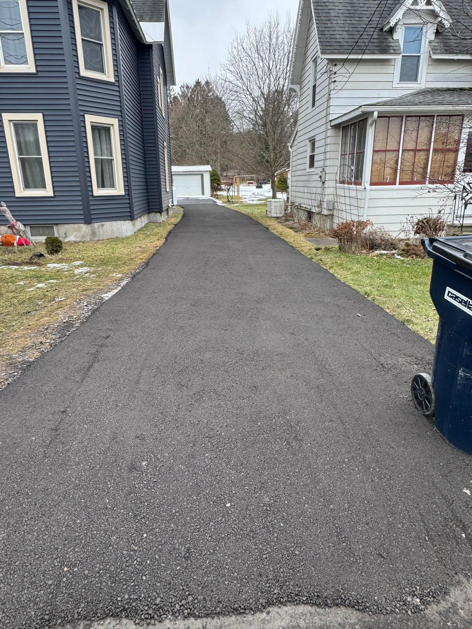 Freshly paved driveway between two houses on a winter day, with a blue trash bin on the right.