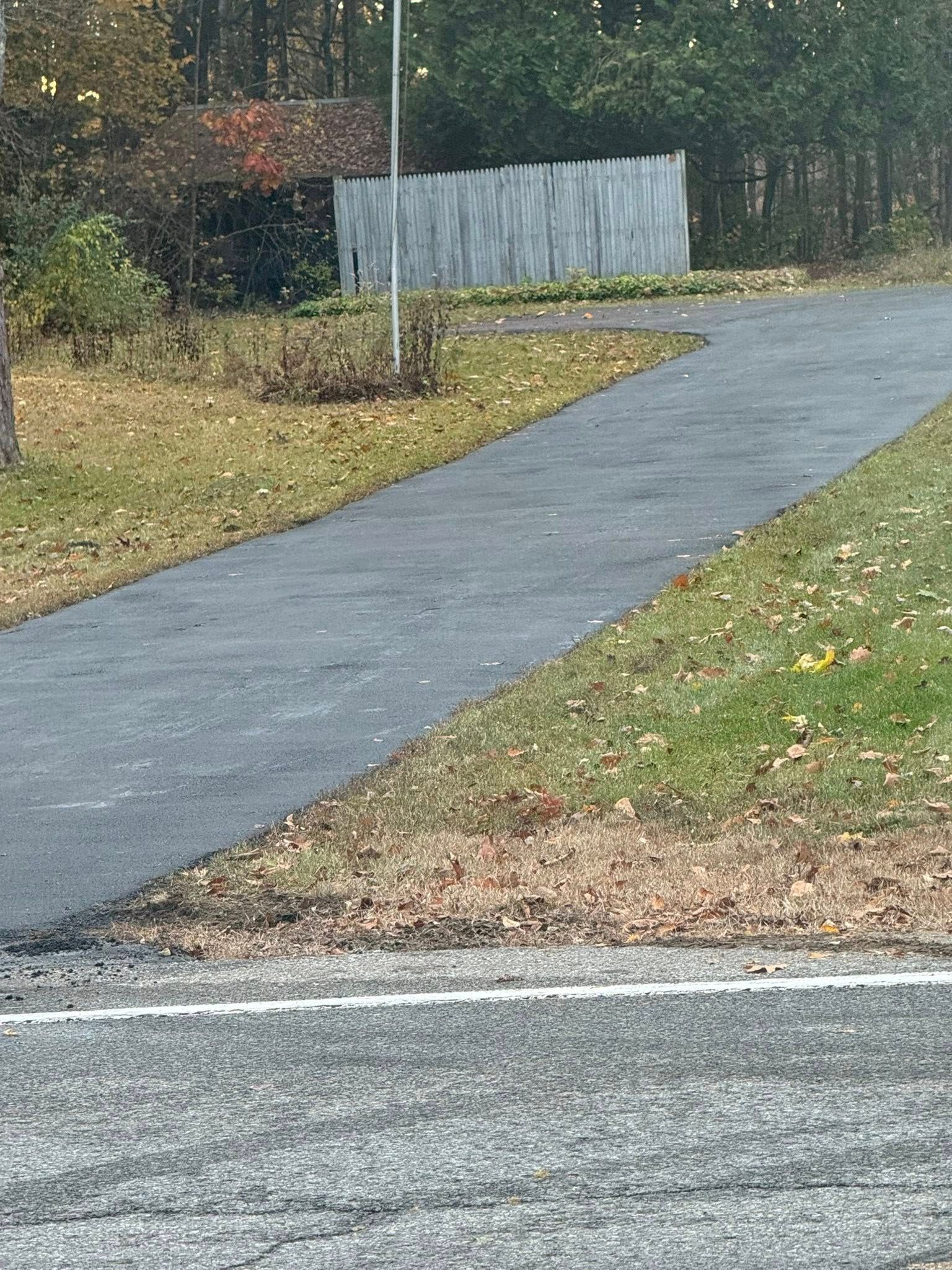 Paved path curving through a grassy area beside a road, with a gray fence and trees in the background.