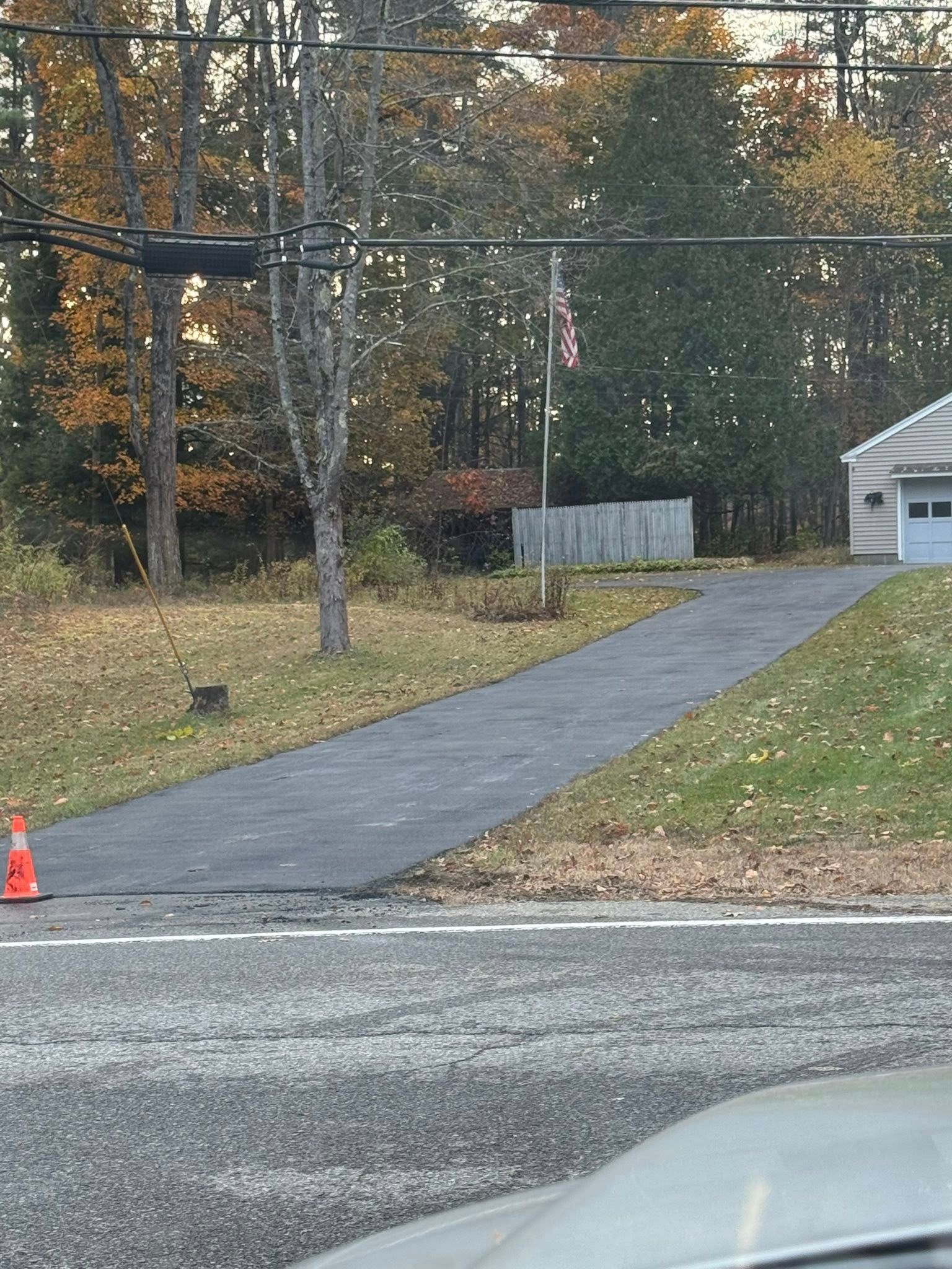 Gravel driveway leading to a white garage beside autumn trees, with a traffic cone at the roadside.