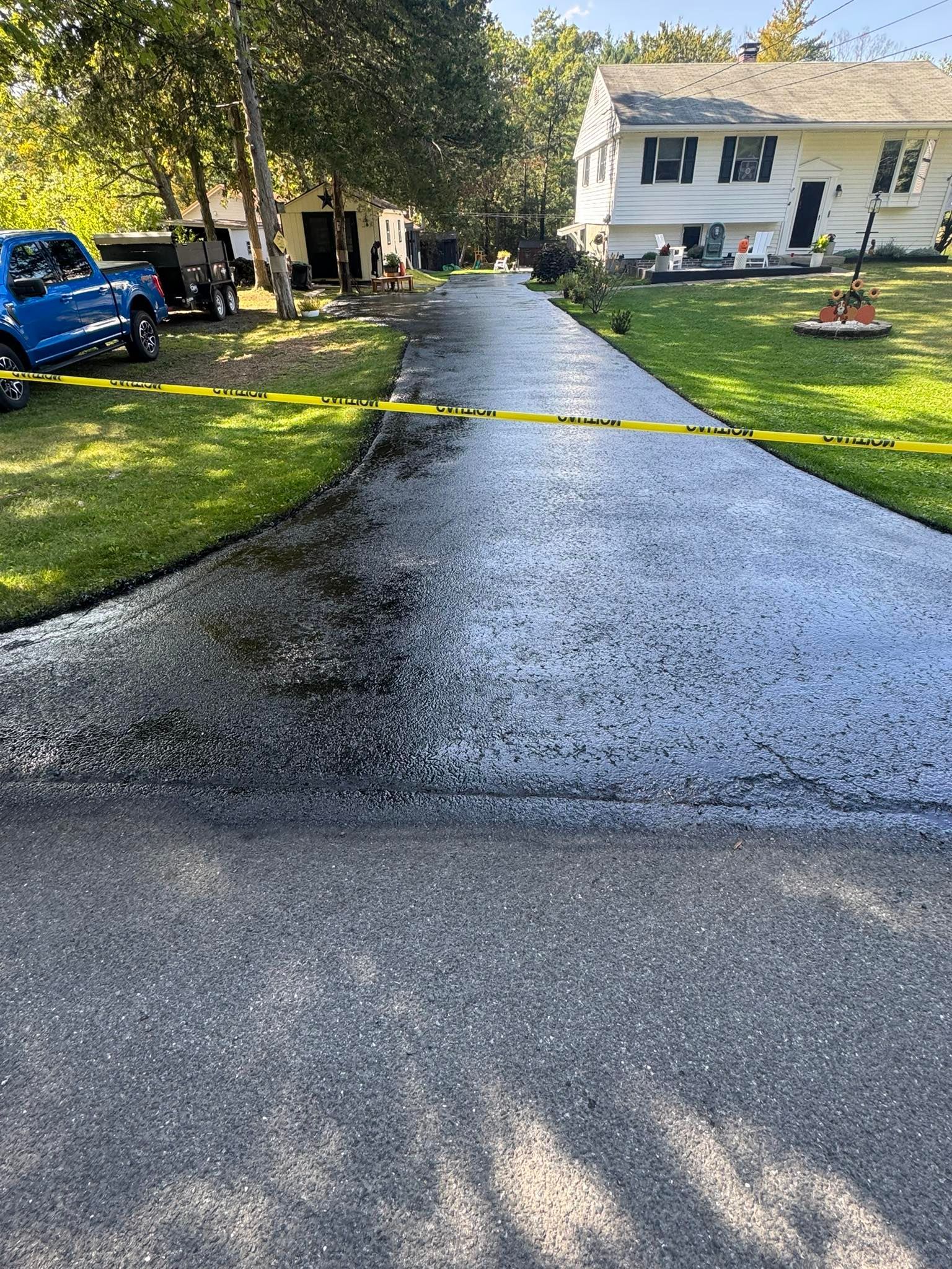 Wet driveway leading to a white house, with yellow caution tape blocking the path and a blue truck on the left.