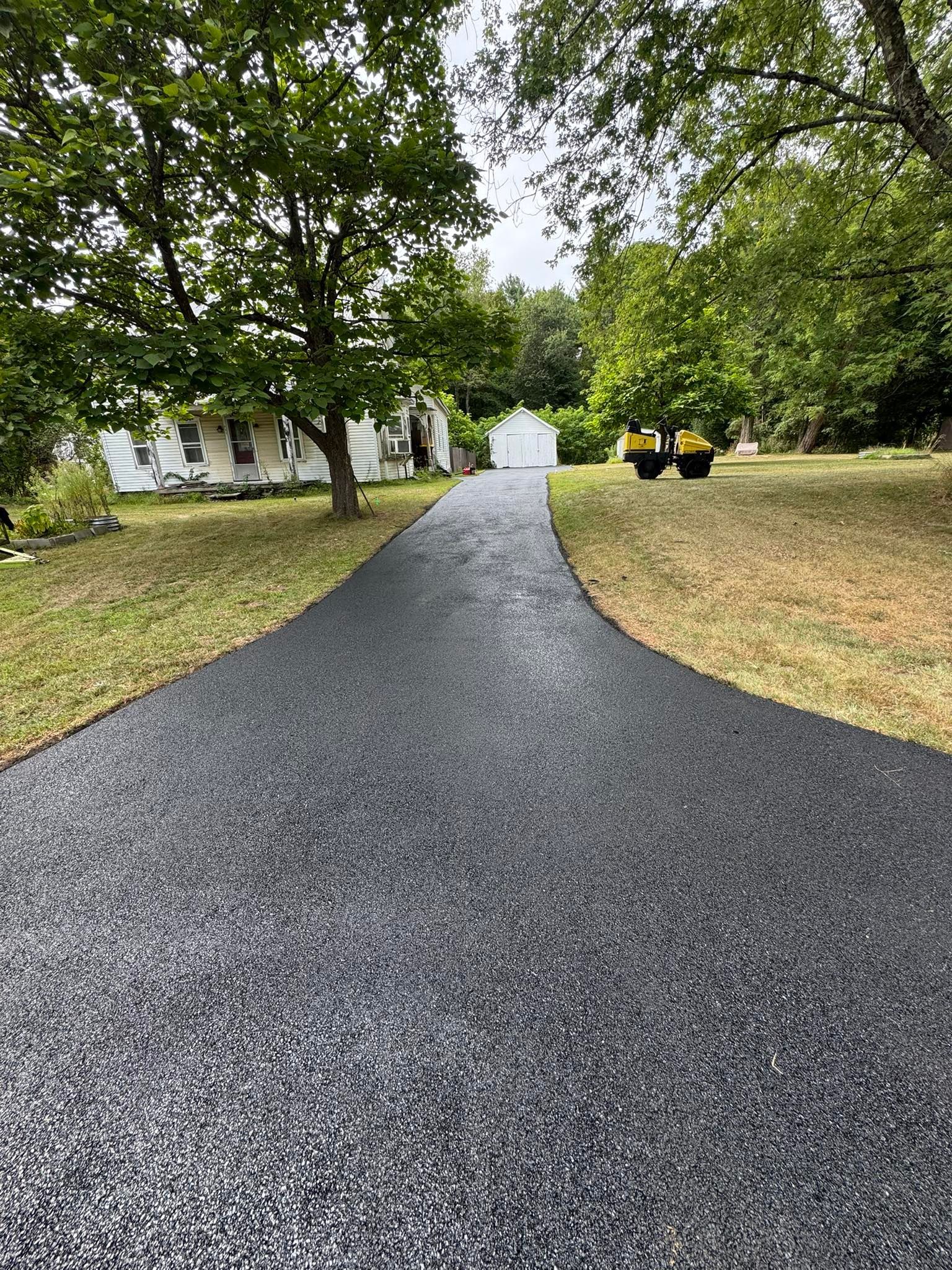 Gravel driveway leading to a white house under large trees on a cloudy day