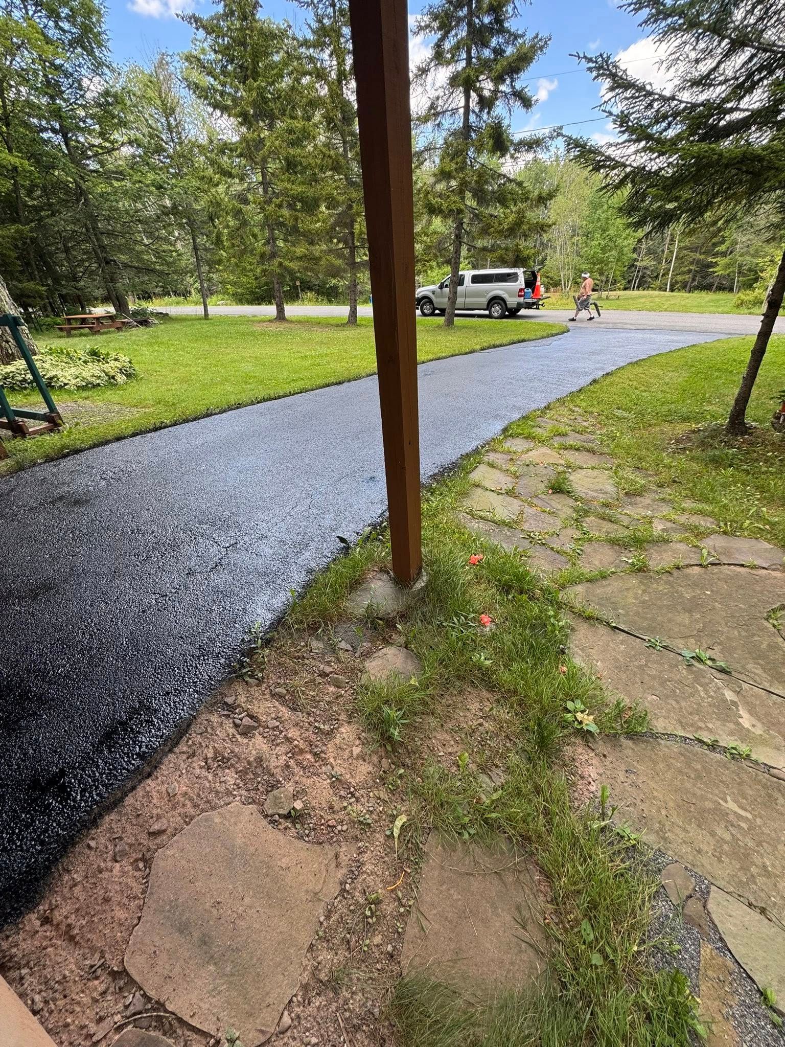 Gravel path under a porch beside a stone patio, leading through a grassy yard with trees and a parked truck.