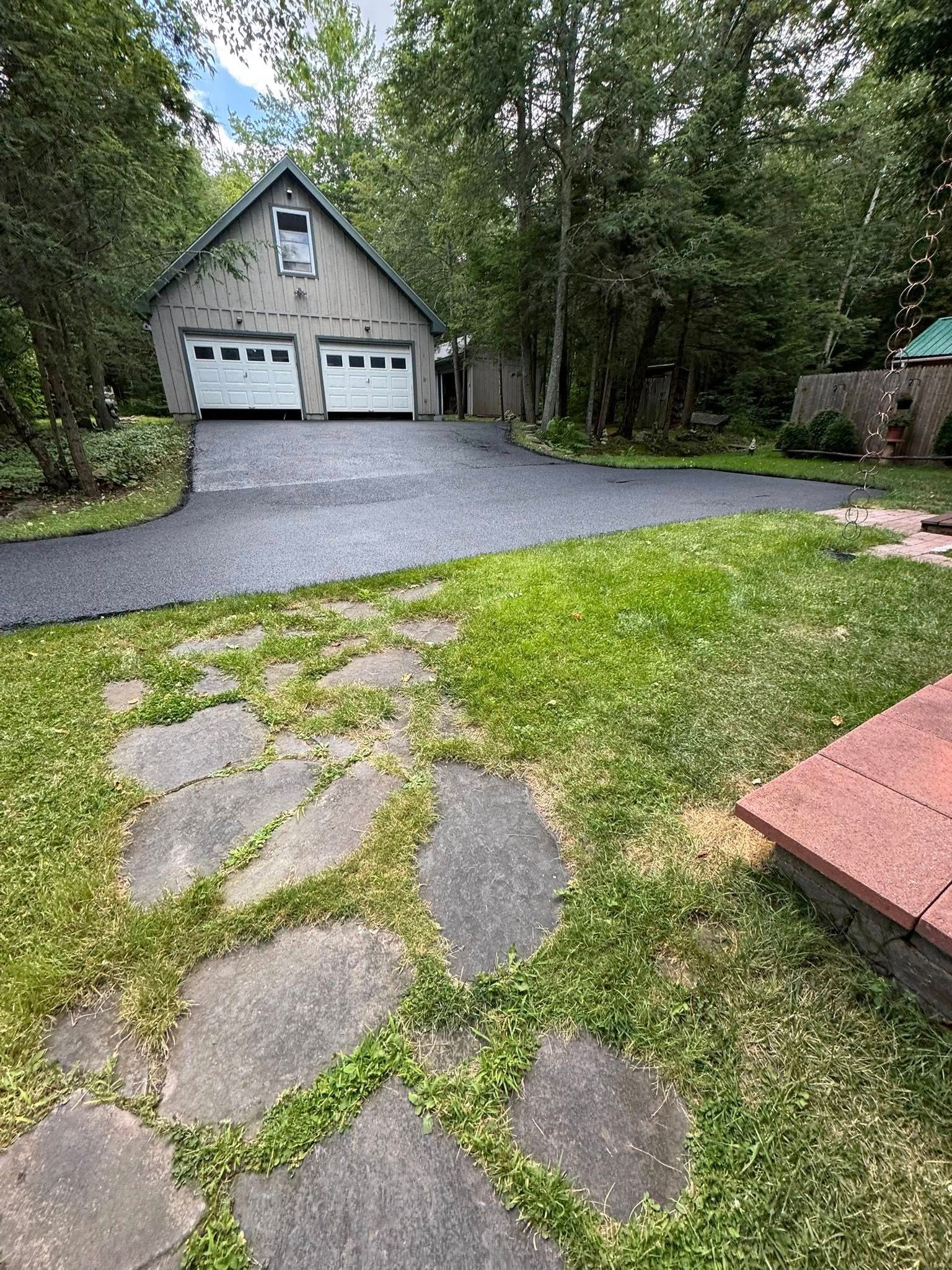 Gravel driveway leading to a gray garage beside a wooded yard with stone stepping path and patio edge