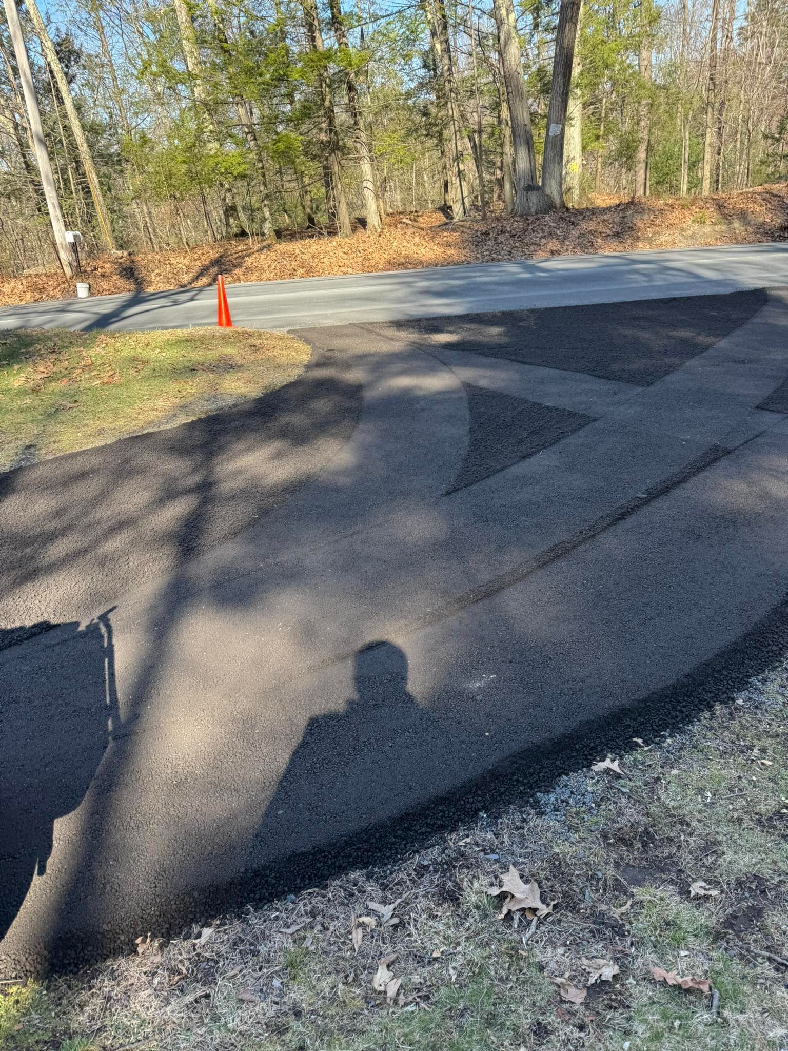 Freshly paved dark asphalt road curve beside woods with an orange cone and leaf litter