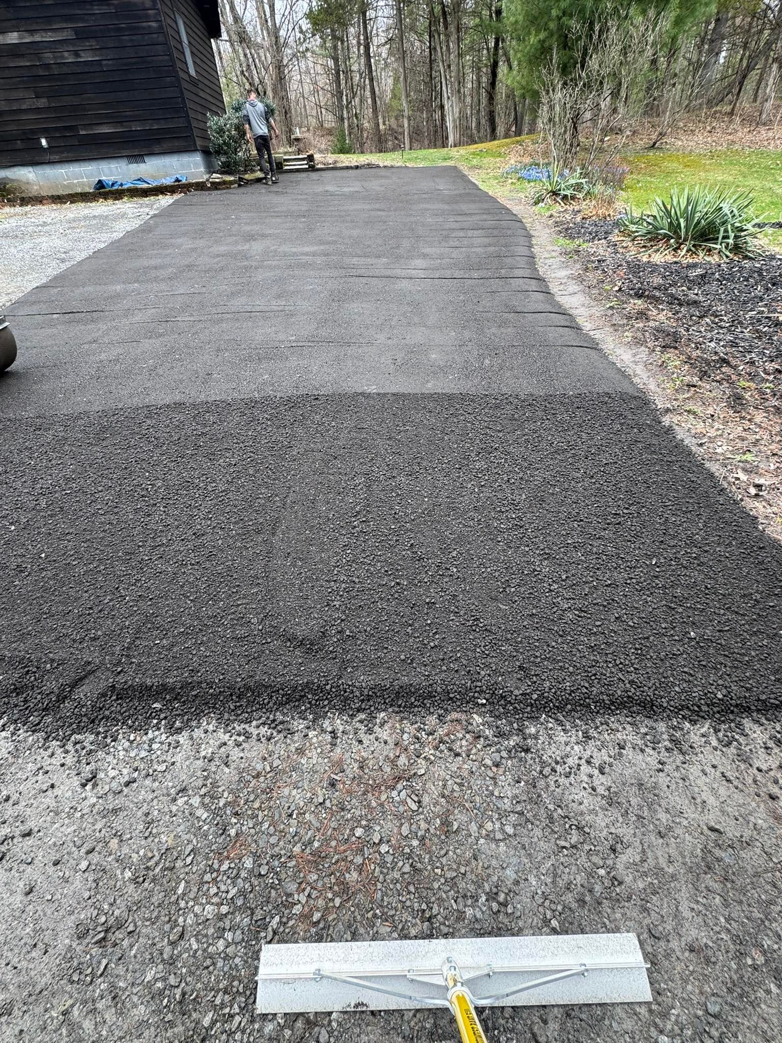 Freshly paved dark gravel path leading into a wooded yard, with a small taped marker in the foreground