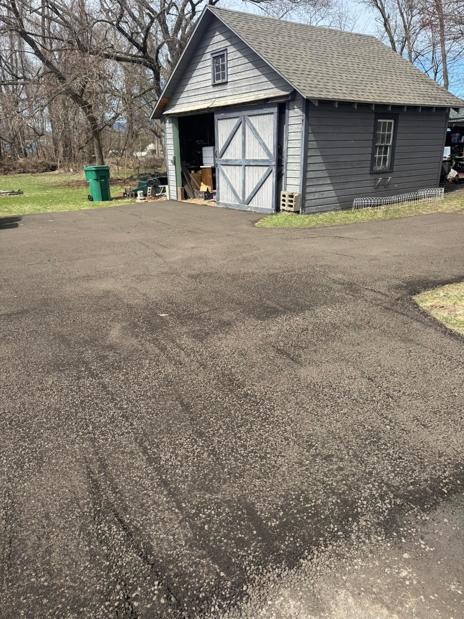 Gravel driveway beside a gray shed with a white double door and green trash bin, in a wooded yard.