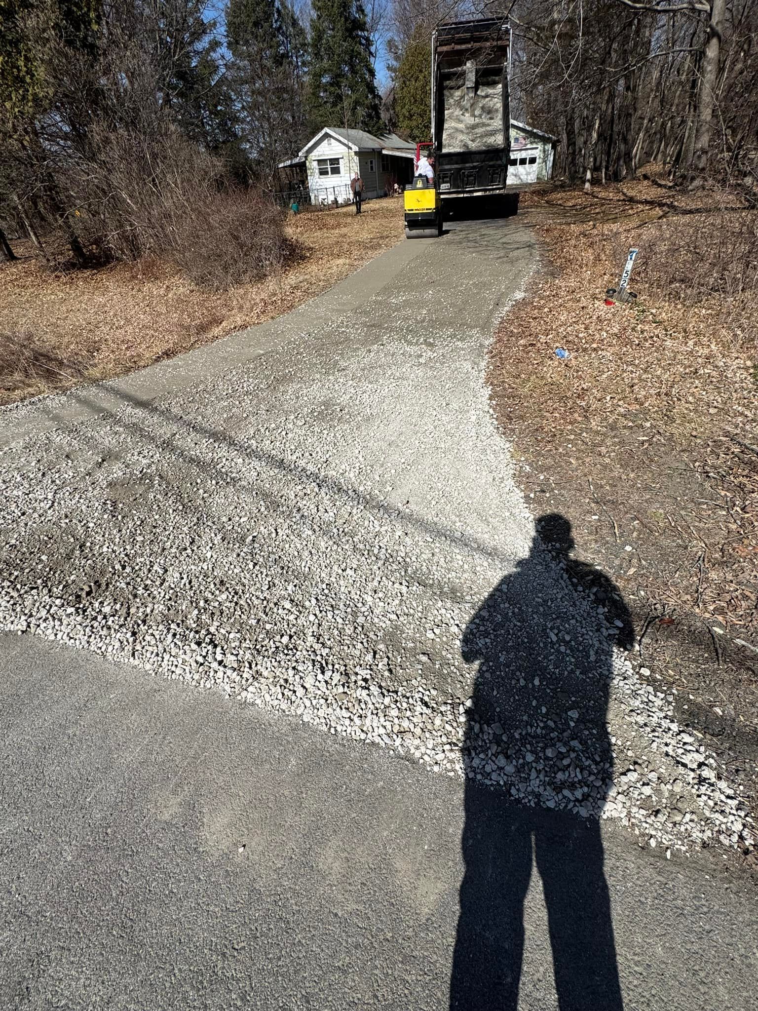 Gravel driveway leading to a house with a car in the distance and a person’s shadow in the foreground