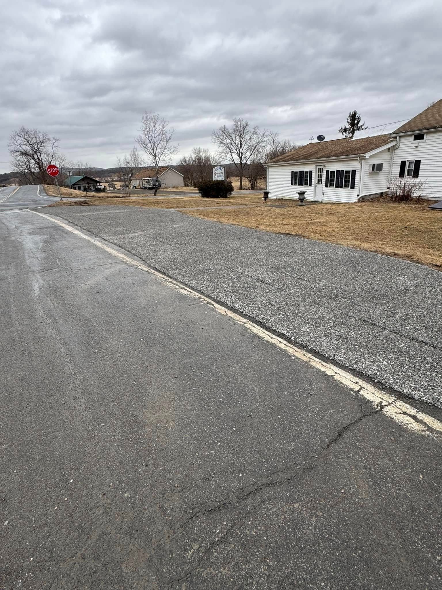 Rural road with gravel shoulder, cloudy sky, and a white house beside a fenced yard