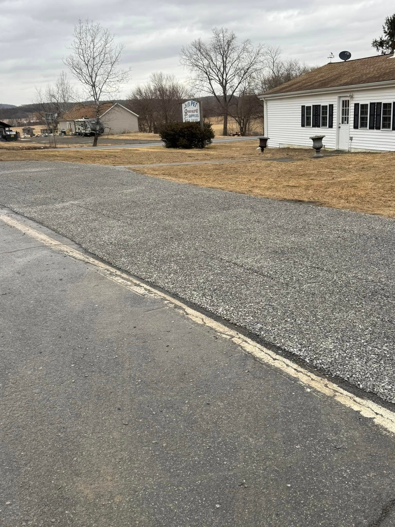 Gravel road beside a white house and leafless trees under a cloudy sky