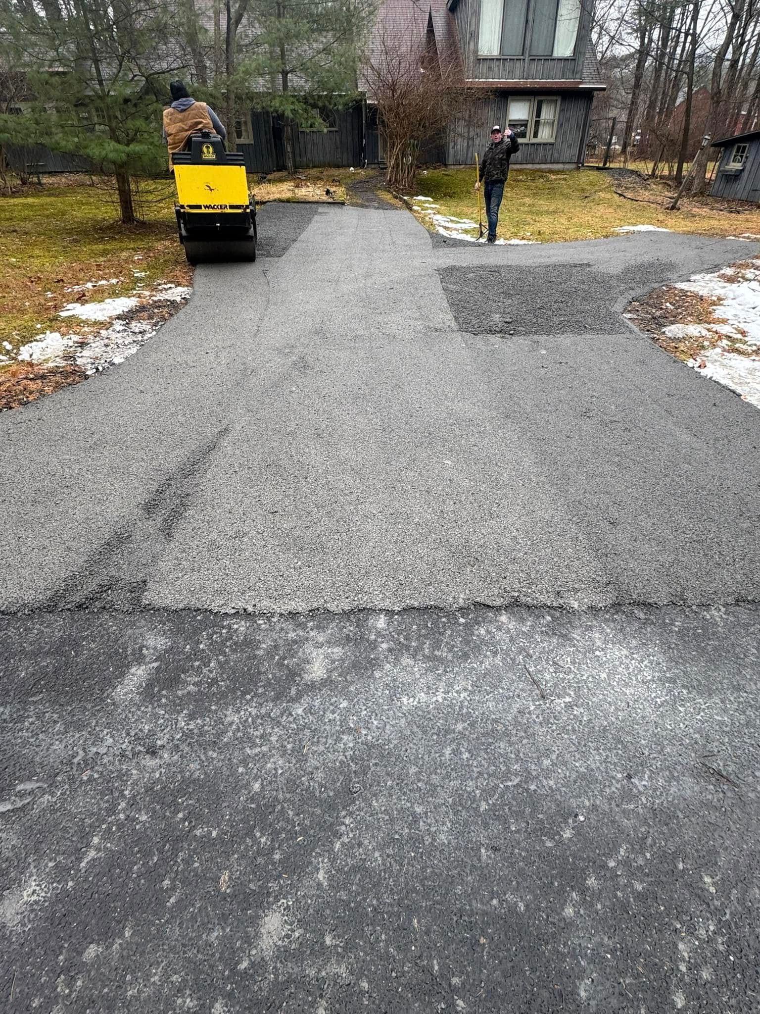 Freshly paved driveway with a yellow road machine and a worker in a wooded residential area