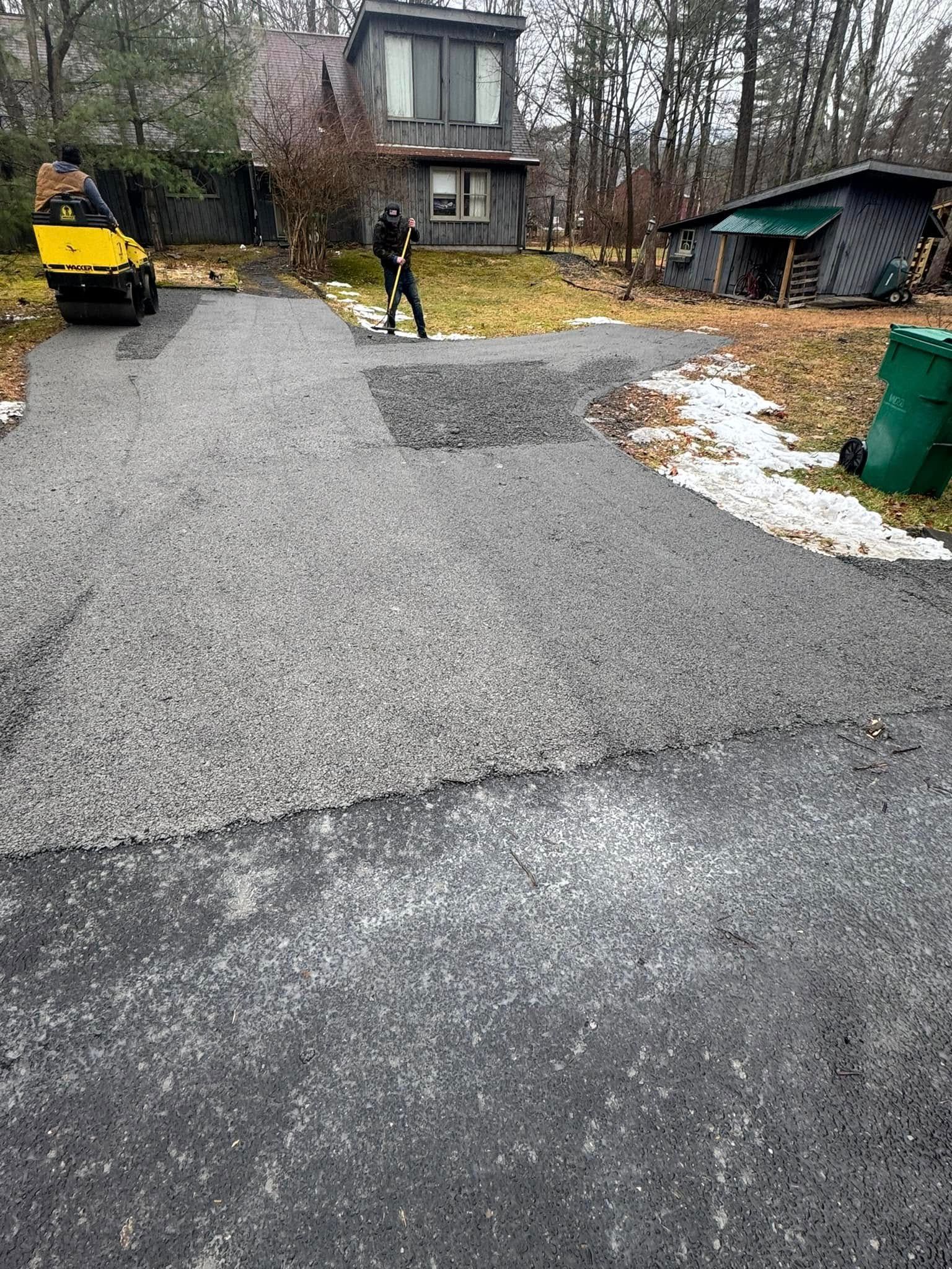 Gravel driveway with a worker in yellow near a house, leaf-covered yard, and a green bin on the right