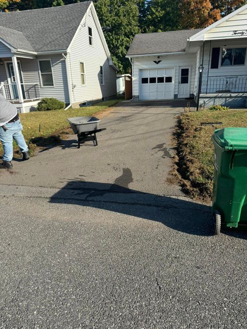 Residential driveway with a wheelbarrow, worker at left, and green trash bin near garage