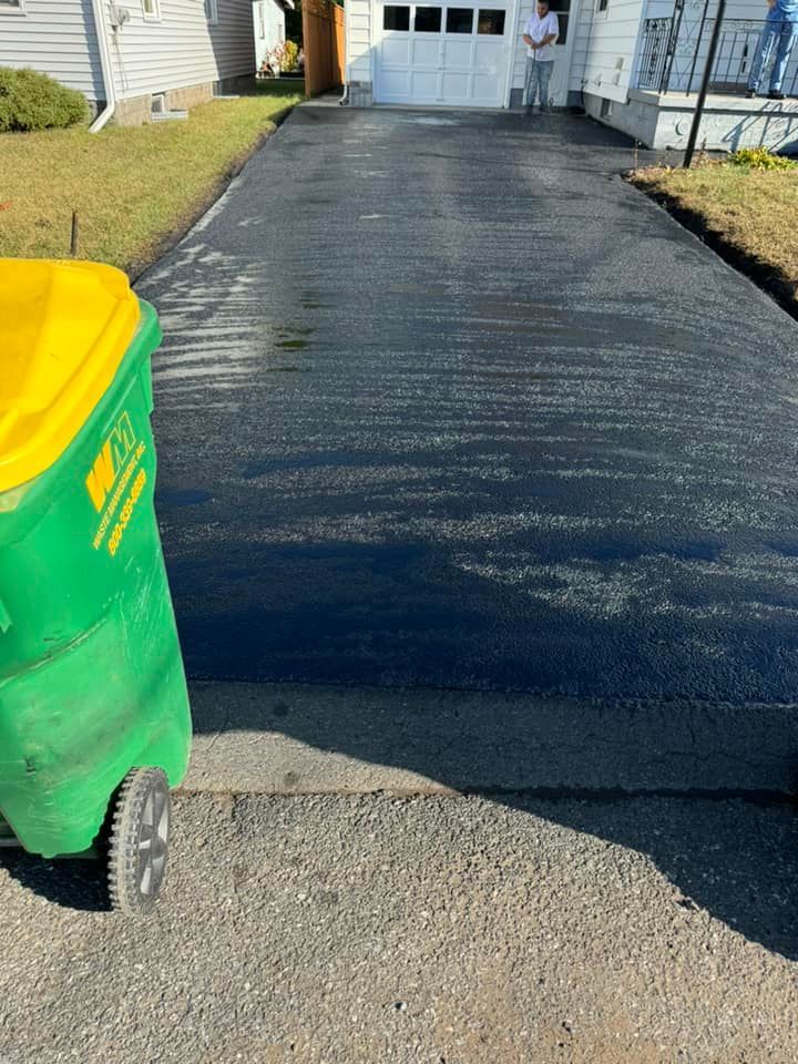 Driveway resurfaced with fresh black asphalt beside a green-and-yellow trash bin.
