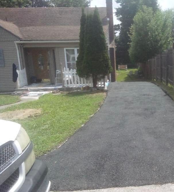 Freshly paved driveway beside a house, with a white fence, front lawn, and parked car in the foreground