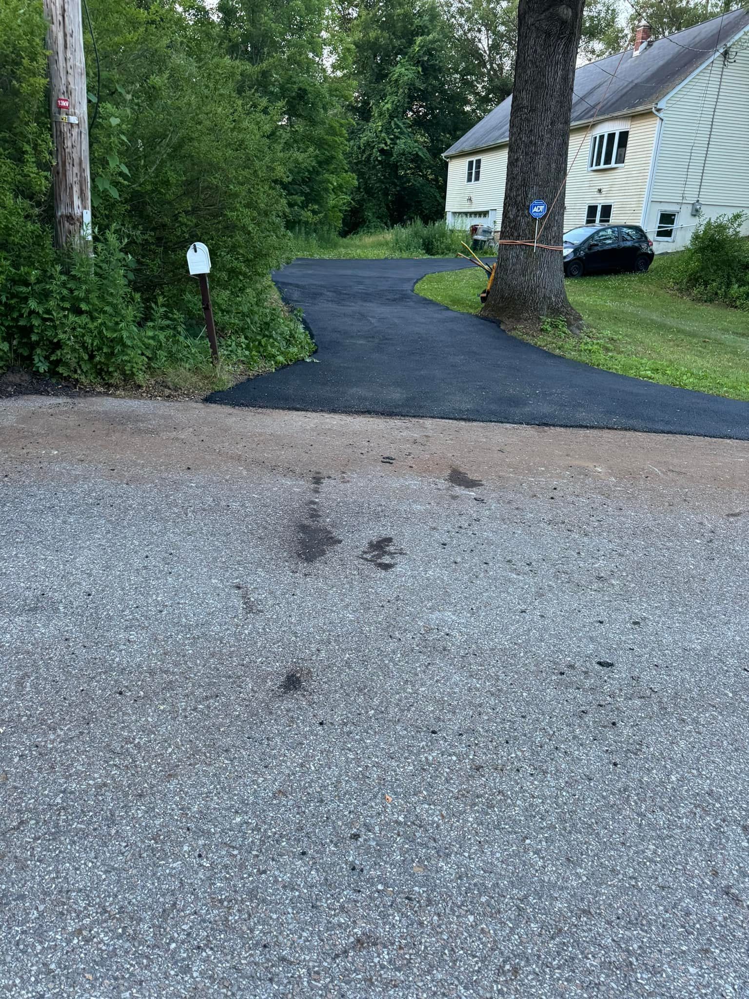 Gravel driveway leading to a white house, bordered by trees and wet asphalt.