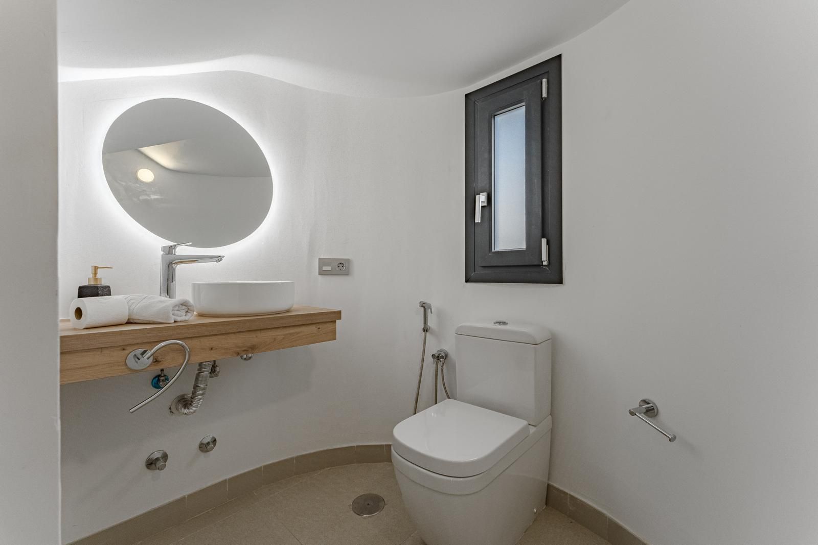 Baño moderno y accesible con lavabo mural, espejo redondo, inodoro y ventana.