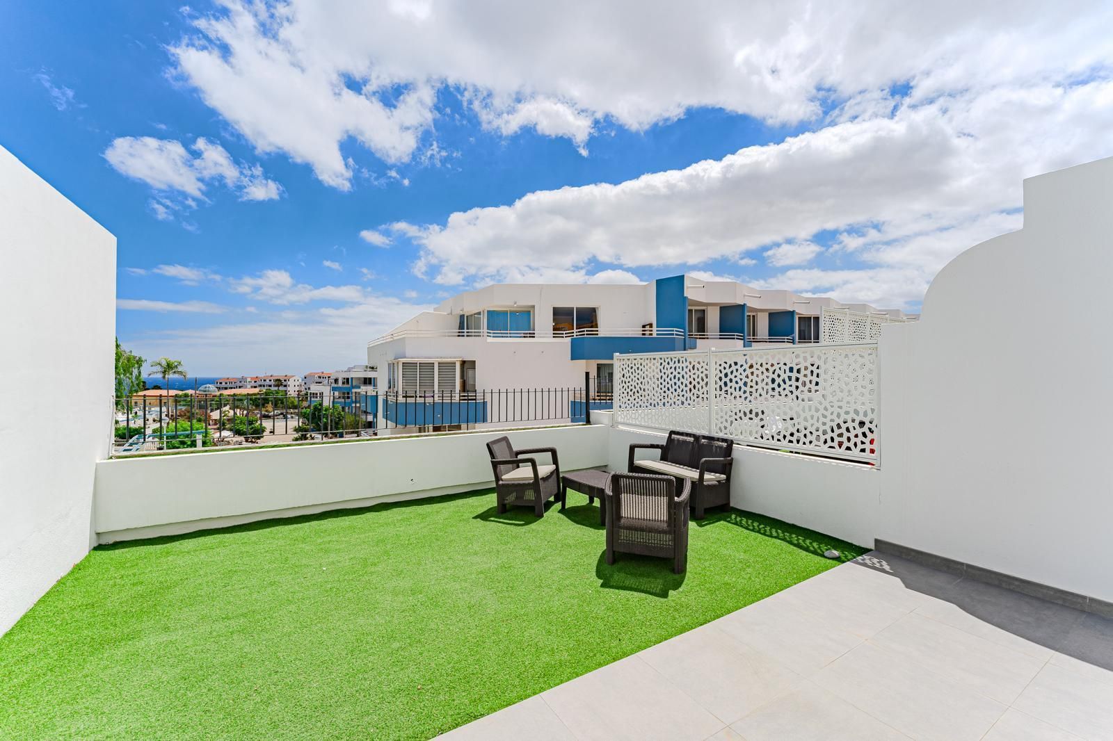 Terraza en la azotea con césped verde, sillas negras y paredes blancas bajo un cielo azul con nubes.