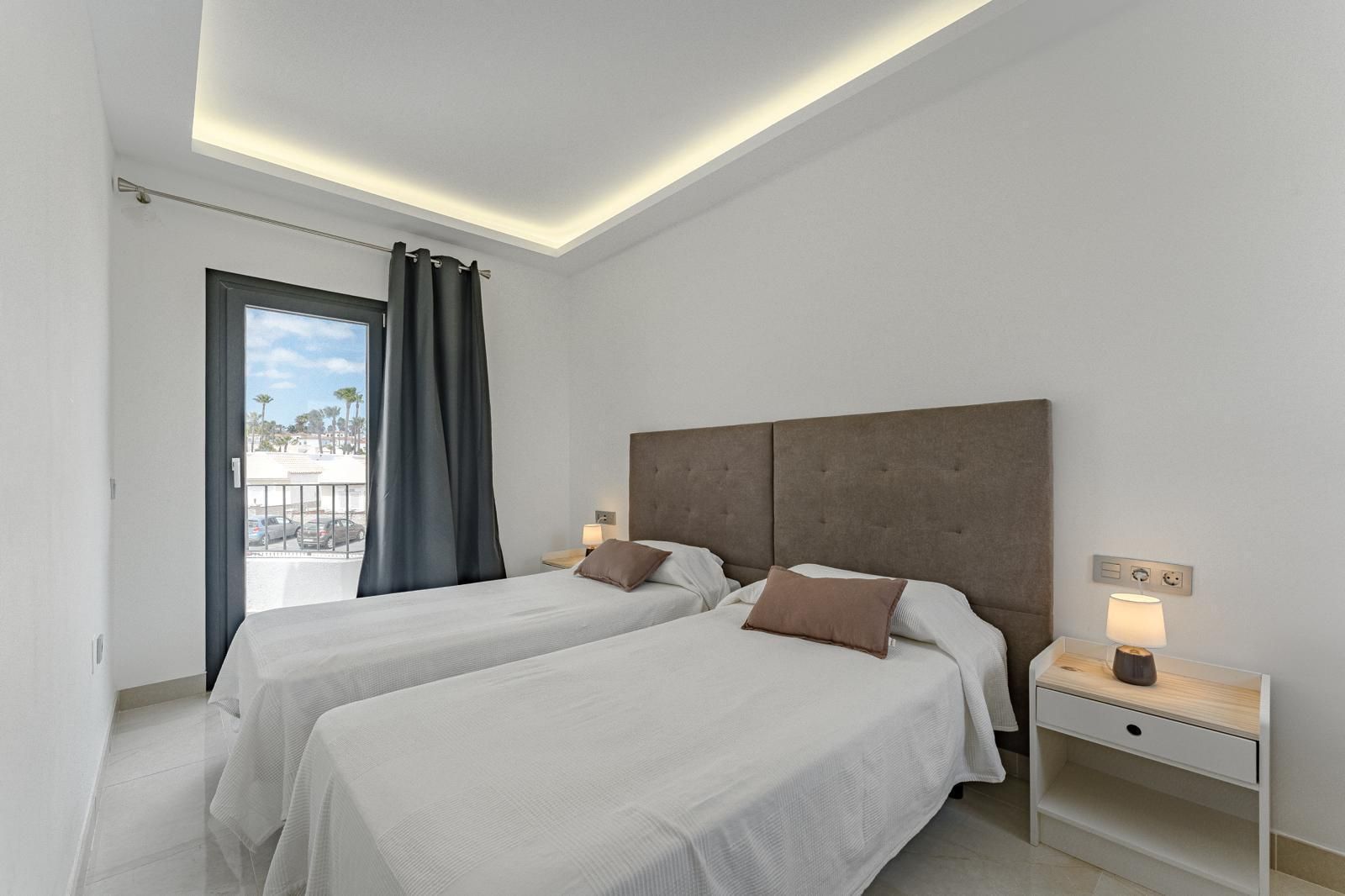 Habitación luminosa y moderna con dos camas individuales, cabecero gris, lámpara de mesilla de noche y ventana con balcón.