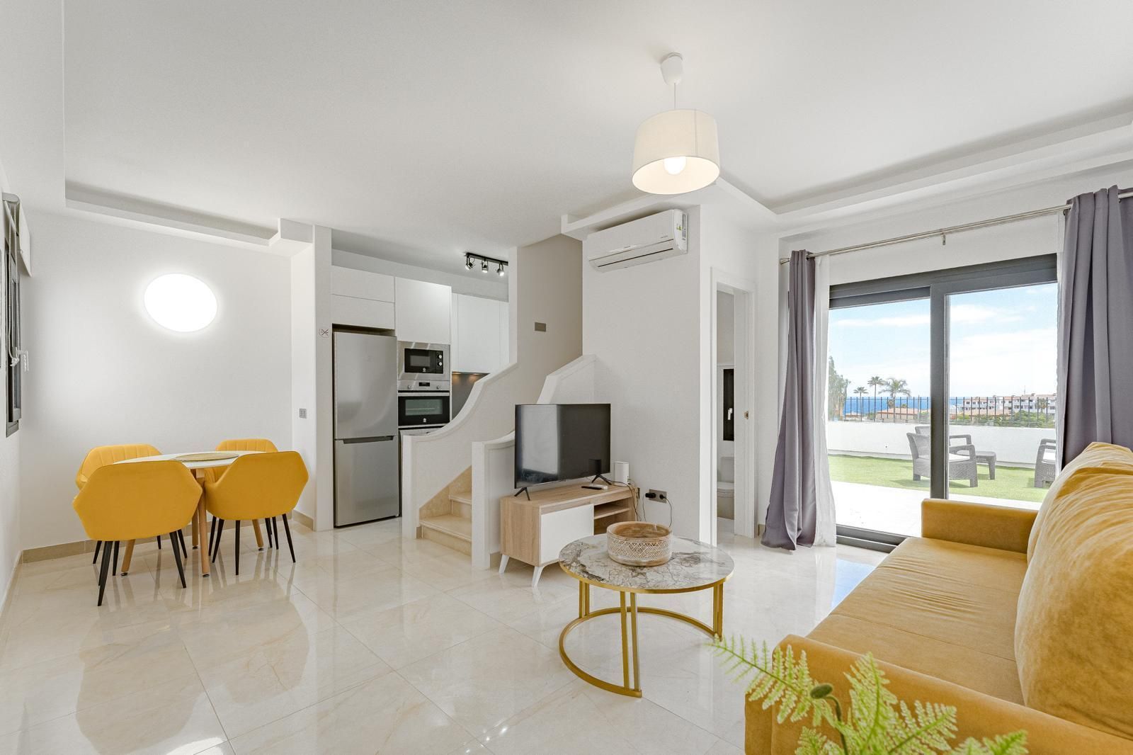 Luminoso salón moderno con sofá amarillo, zona de comedor y puertas correderas de cristal que dan a un patio.