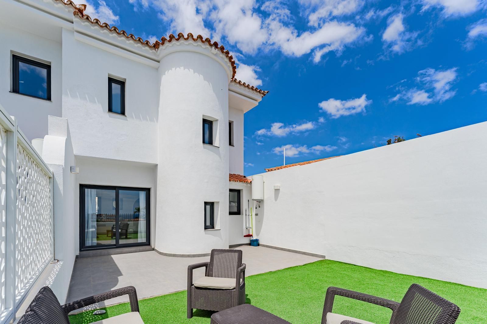 Patio de estuco blanco con césped verde, sillas negras y una puerta corrediza de vidrio bajo un cielo azul.
