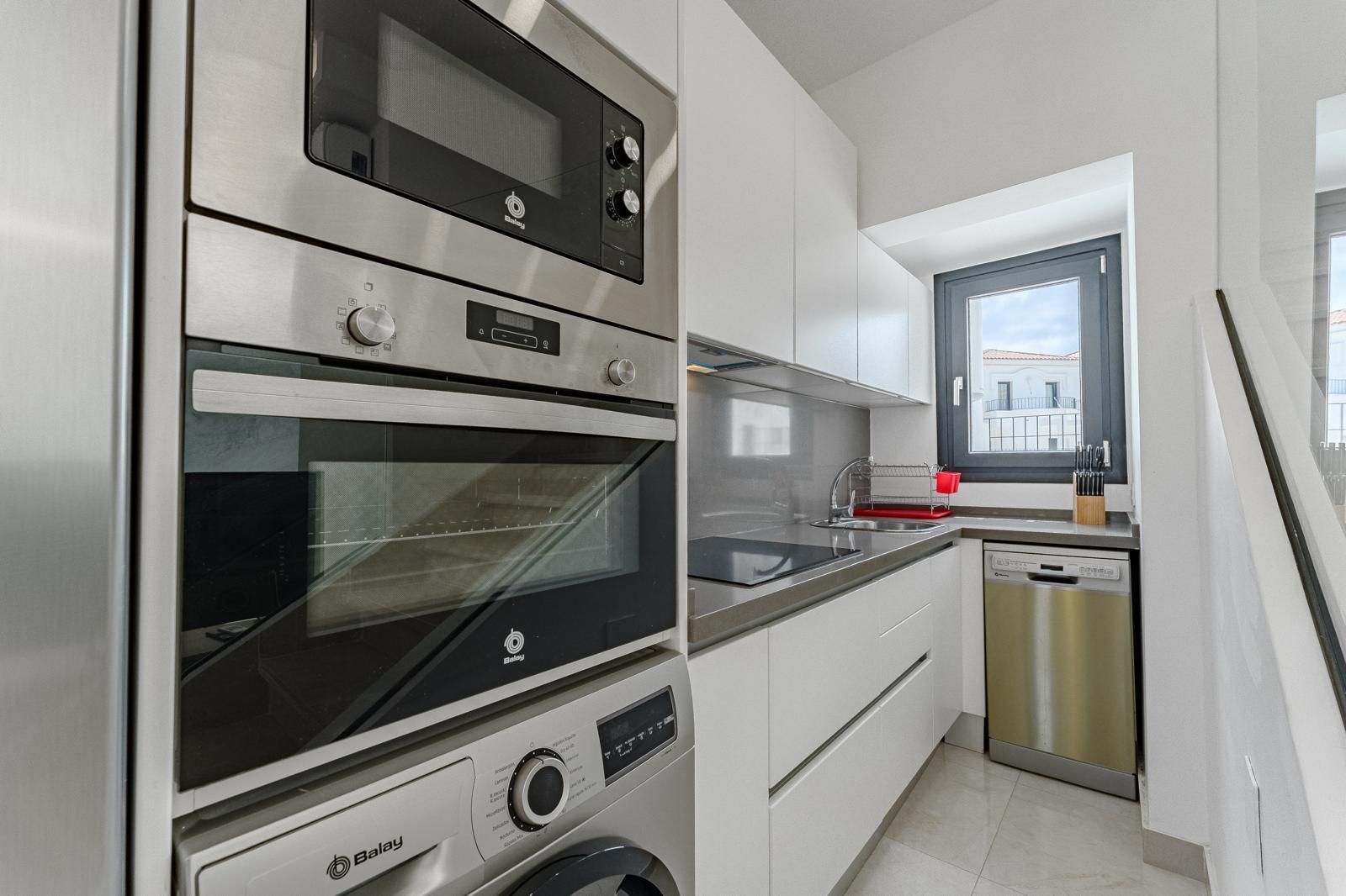 Cocina moderna de color blanco con horno empotrado, microondas, lavavajillas y una ventana sobre el fregadero.