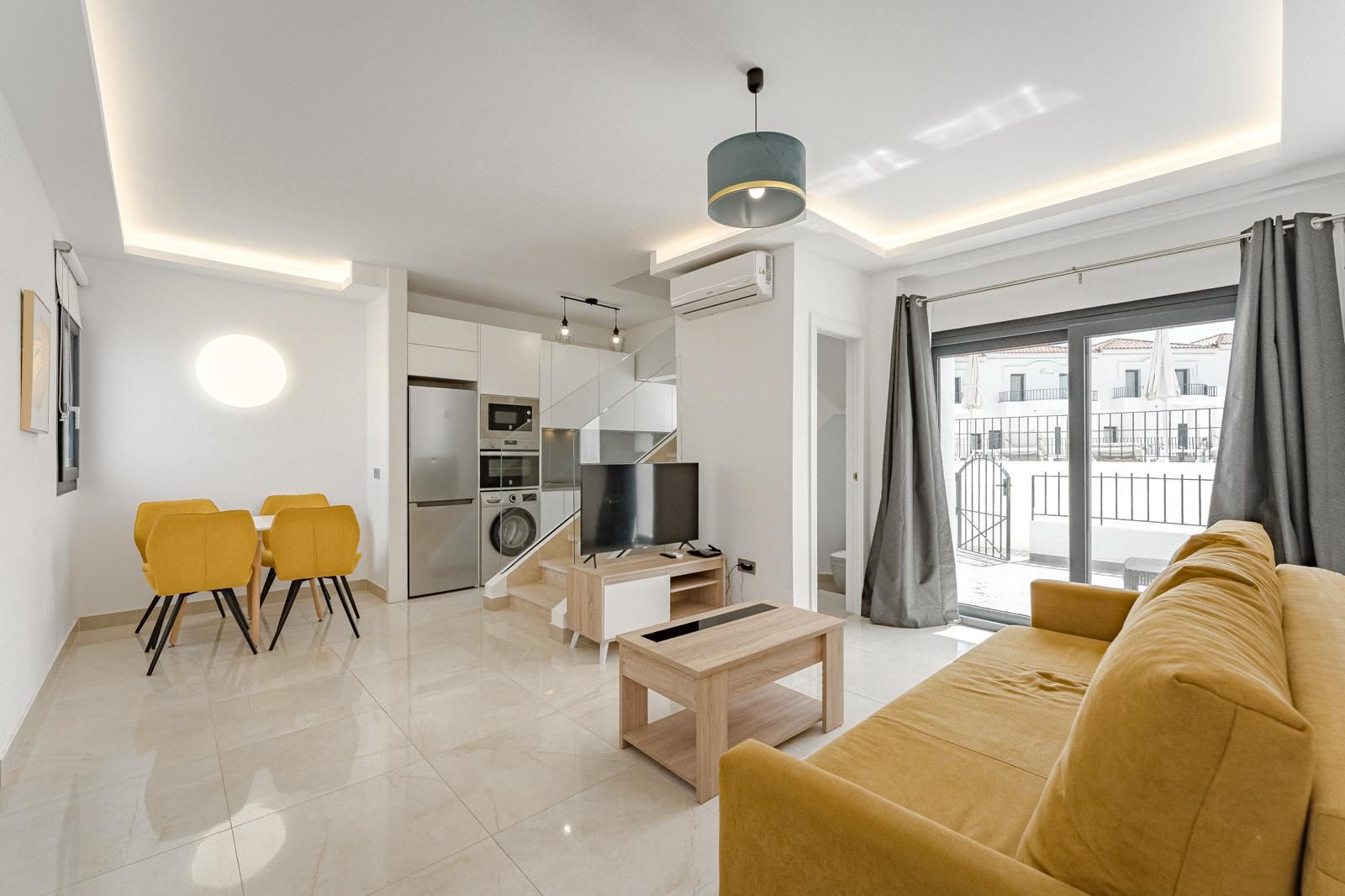 Sala moderna y luminosa con sofá mostaza, comedor, TV y puertas al balcón con cortinas grises.