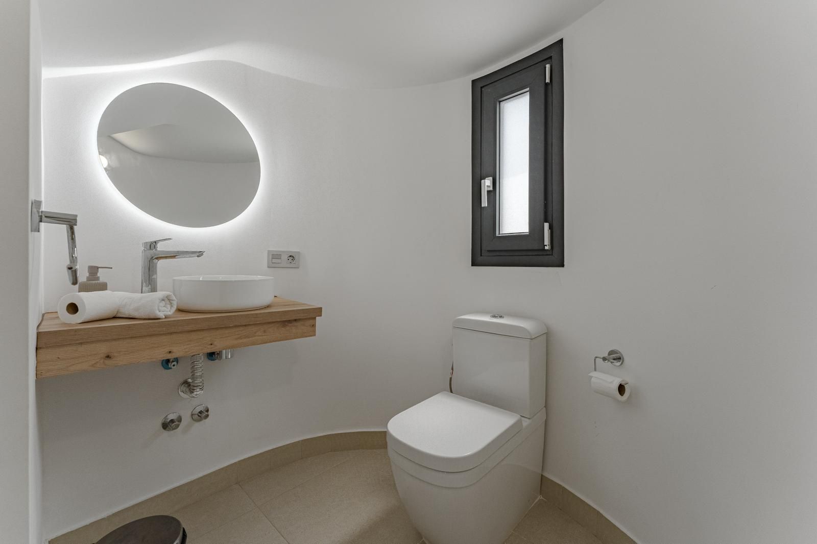Baño minimalista blanco con espejo redondo, lavabo de madera, inodoro y ventana pequeña.