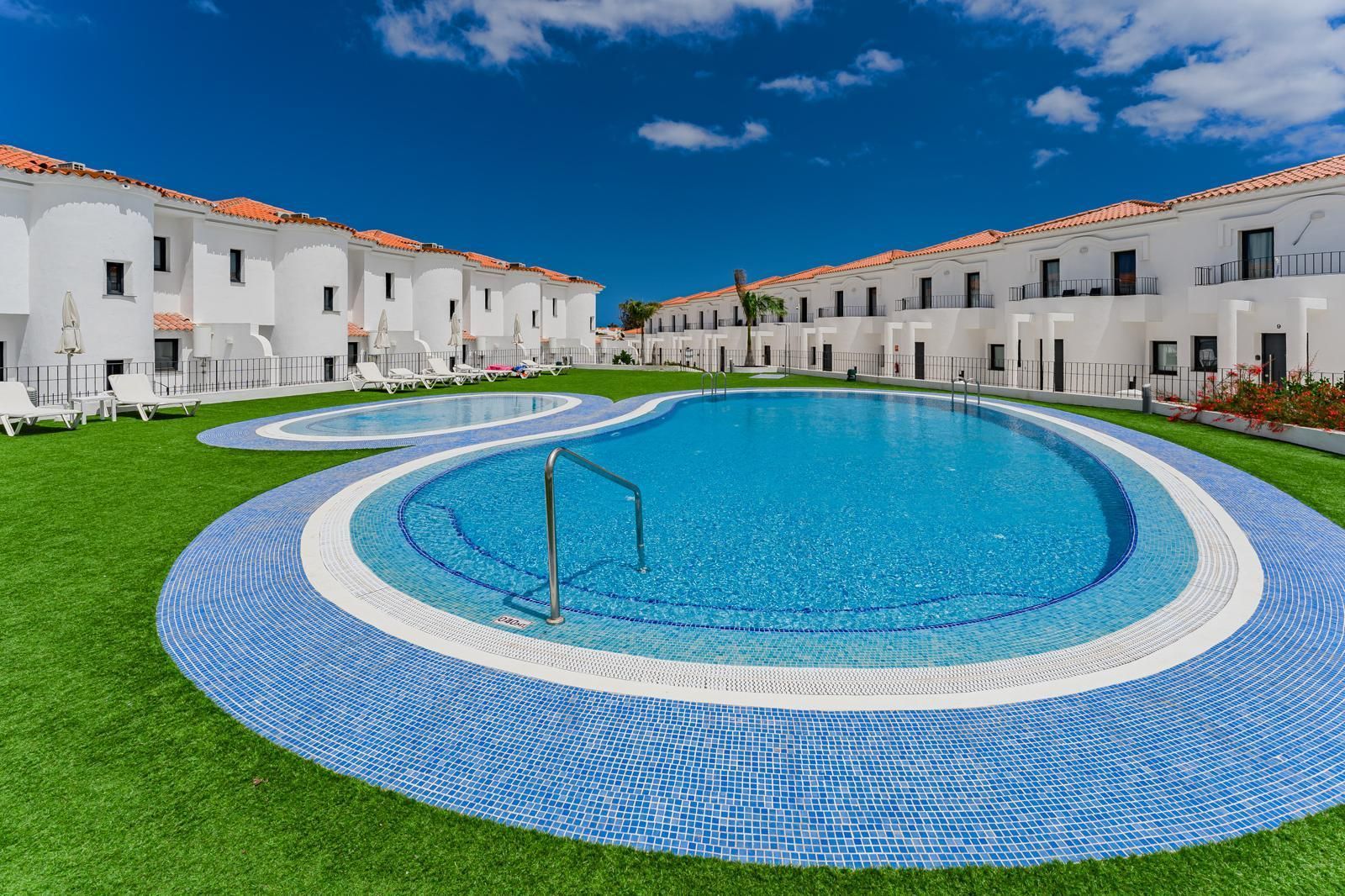 Villas blancas modernas, piscina circular azul con césped verde bajo un cielo despejado.