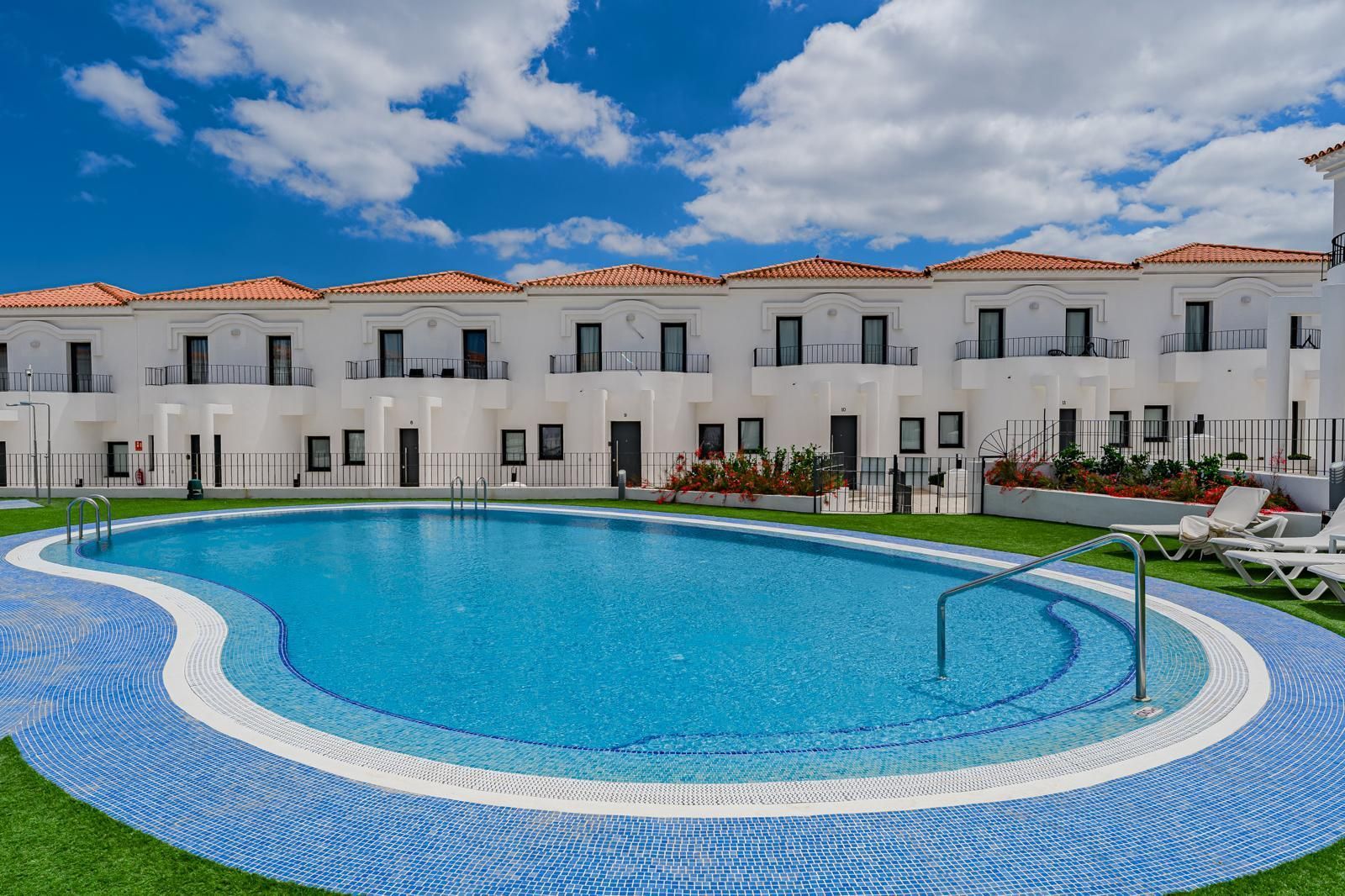 Piscina azul curva frente a un edificio blanco del complejo turístico bajo un cielo brillante.