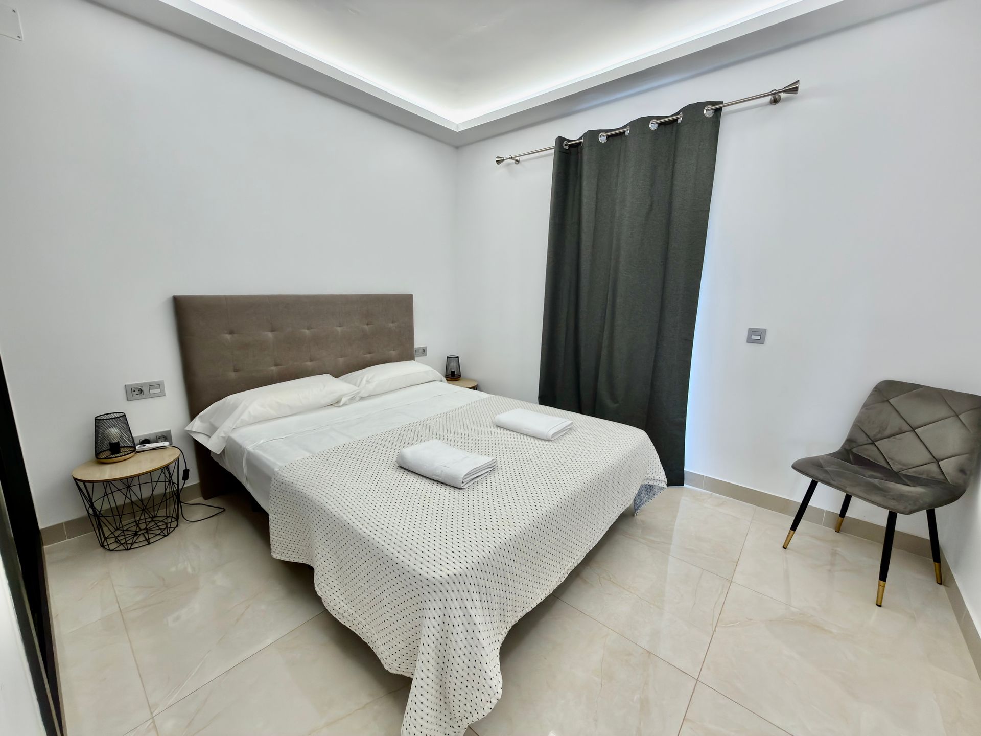 Dormitorio minimalista con una cama, una mesita de noche, cortinas oscuras y una silla sobre un suelo de baldosas.